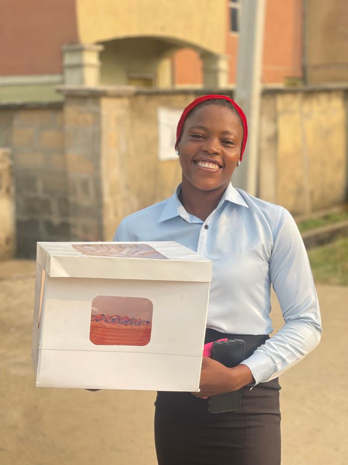 A cheerful woman with a cake box standing outdoors, dressed in a blue shirt and red headband.